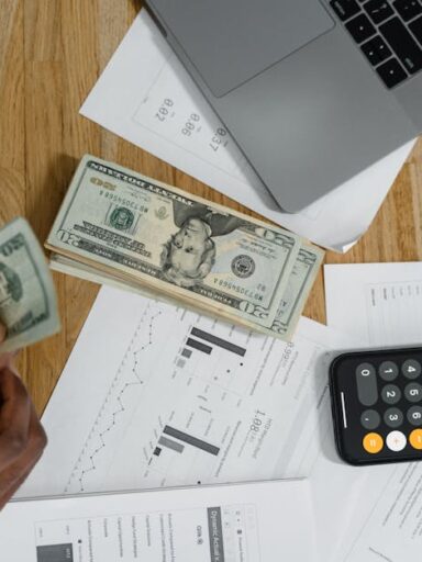 Osoba liczy banknoty dolarowe przy biurku z laptopem, kalkulatorem i wykresami.
