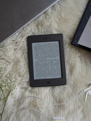 Czytnik e-booków, laptop, otwarta książka i kwiaty na futrzanym kocu.