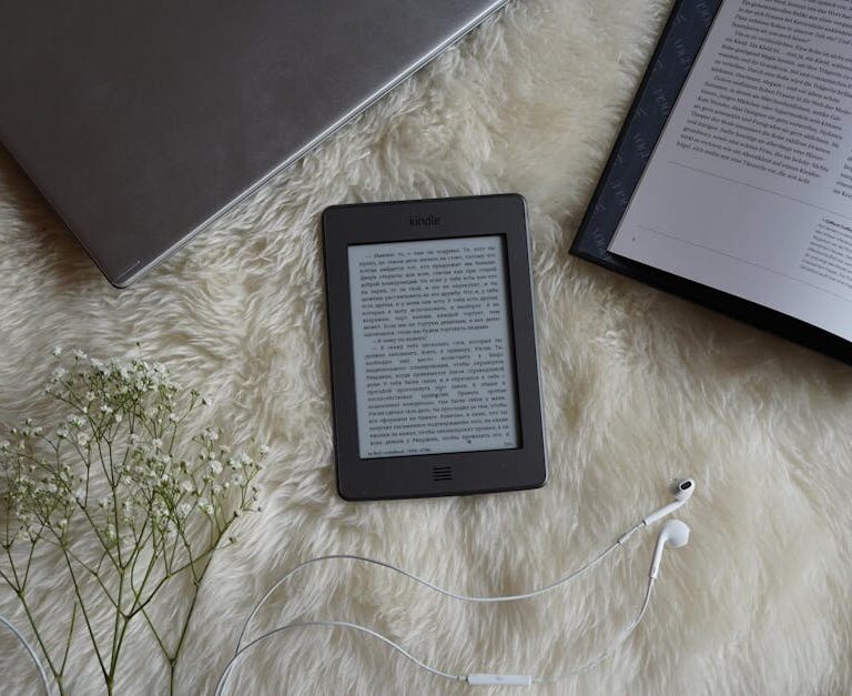 Czytnik e-booków, laptop, otwarta książka i kwiaty na futrzanym kocu.