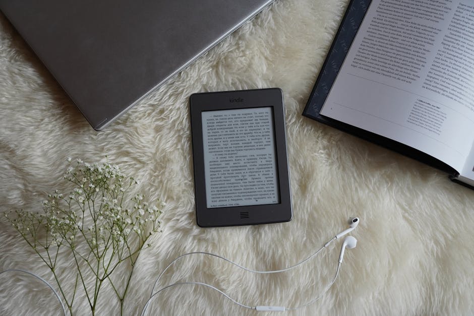 Czytnik e-booków, laptop, otwarta książka i kwiaty na futrzanym kocu.