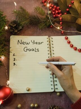 Ręka z długopisem nad otwartym notatnikiem z napisem New Year Goals.
