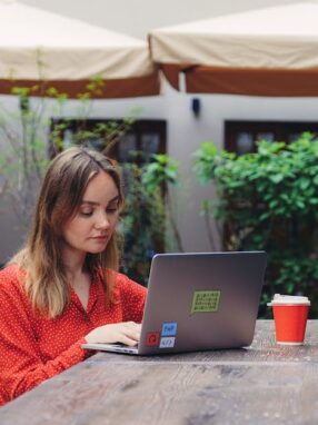 Kobieta w czerwonej bluzce pracuje przy laptopie w kawiarnianym ogródku.