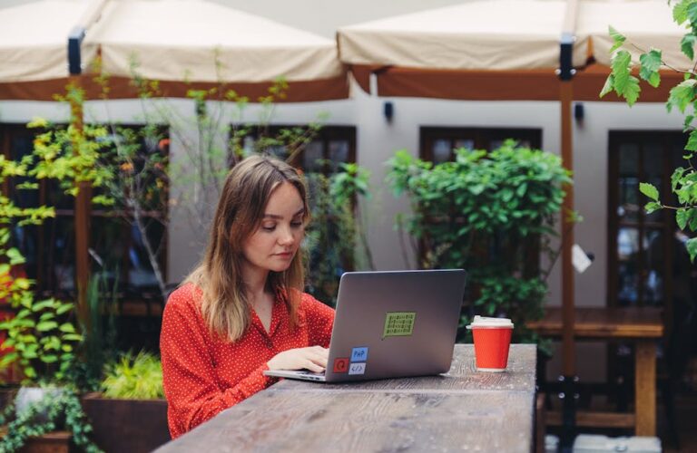 Kobieta w czerwonej bluzce pracuje przy laptopie w kawiarnianym ogródku.