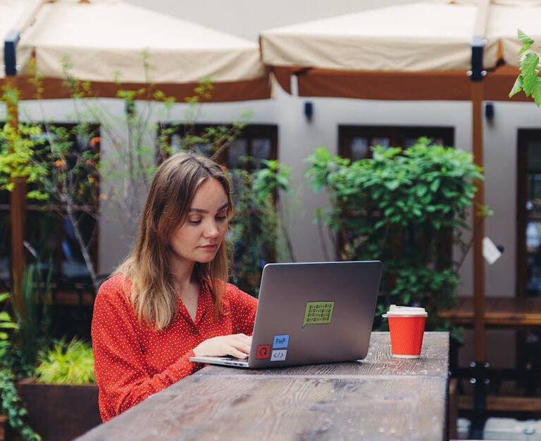 Kobieta w czerwonej bluzce pracuje przy laptopie w kawiarnianym ogródku.