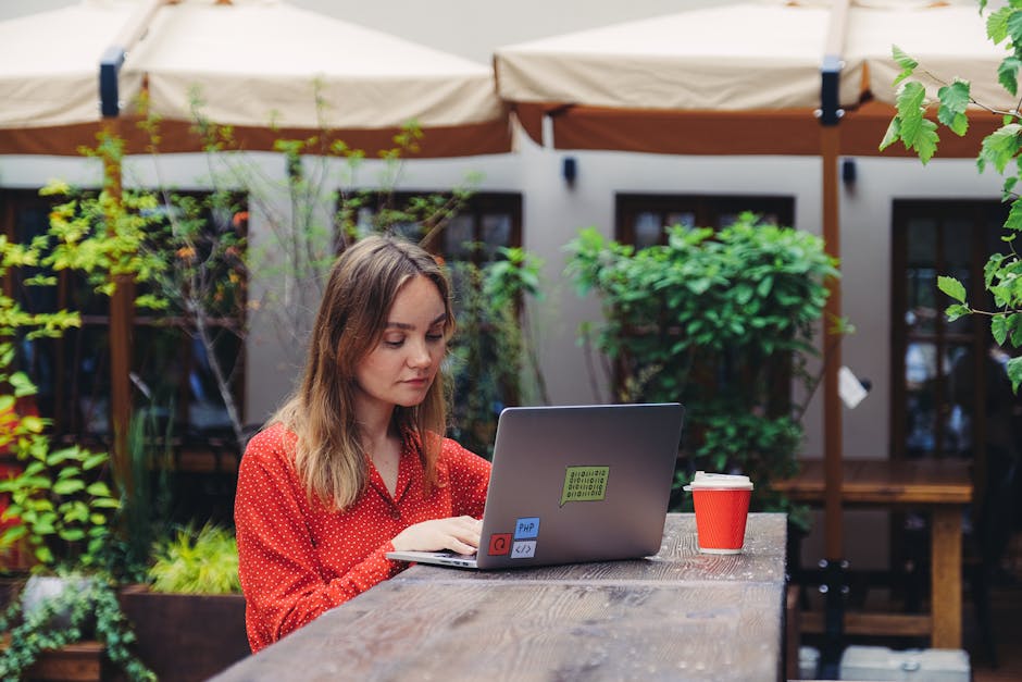 Kobieta w czerwonej bluzce pracuje przy laptopie w kawiarnianym ogródku.