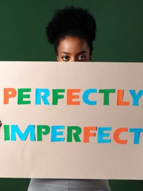 Kobieta trzymająca przed twarzą tekturową tabliczkę z napisem „PERFECTLY IMPERFECT”.