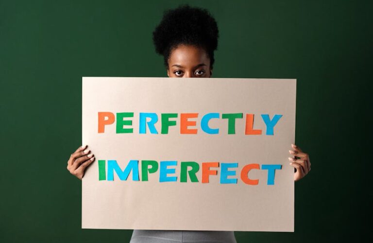 Kobieta trzymająca przed twarzą tekturową tabliczkę z napisem „PERFECTLY IMPERFECT”.