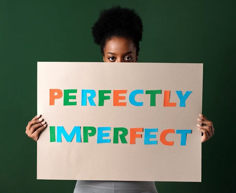 Kobieta trzymająca przed twarzą tekturową tabliczkę z napisem „PERFECTLY IMPERFECT”.