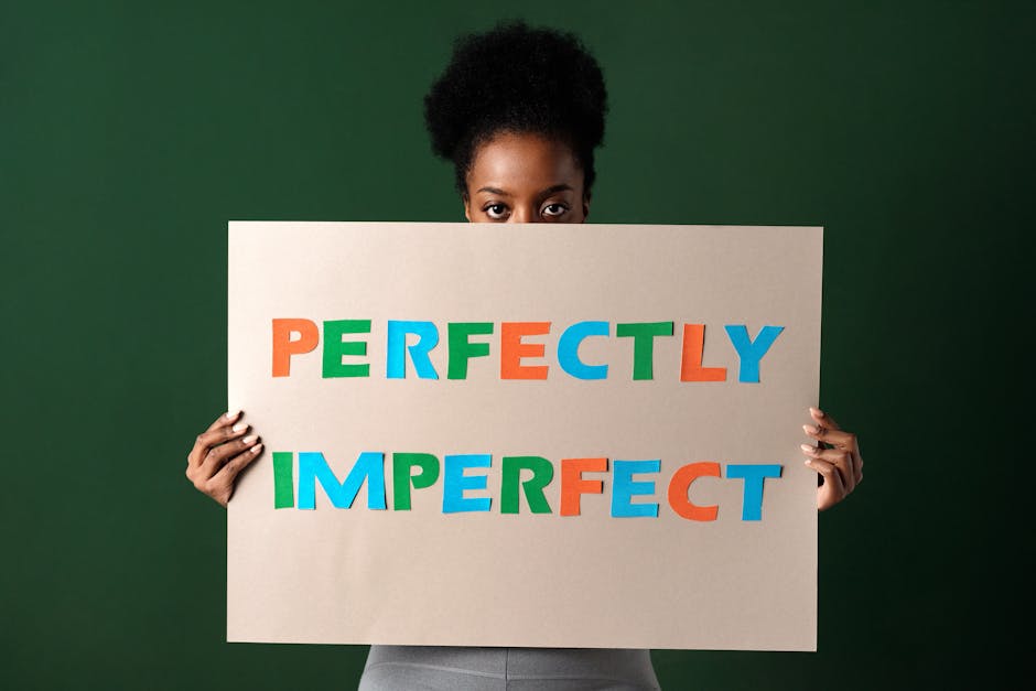 Kobieta trzymająca przed twarzą tekturową tabliczkę z napisem „PERFECTLY IMPERFECT”.