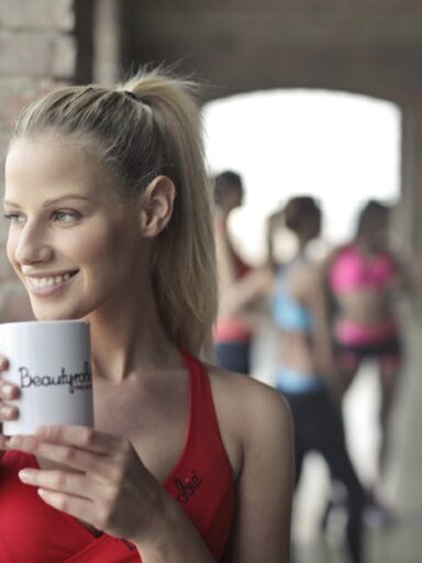 Kobieta w czerwonym topie trzyma biały kubek przy oknie w sali fitness.