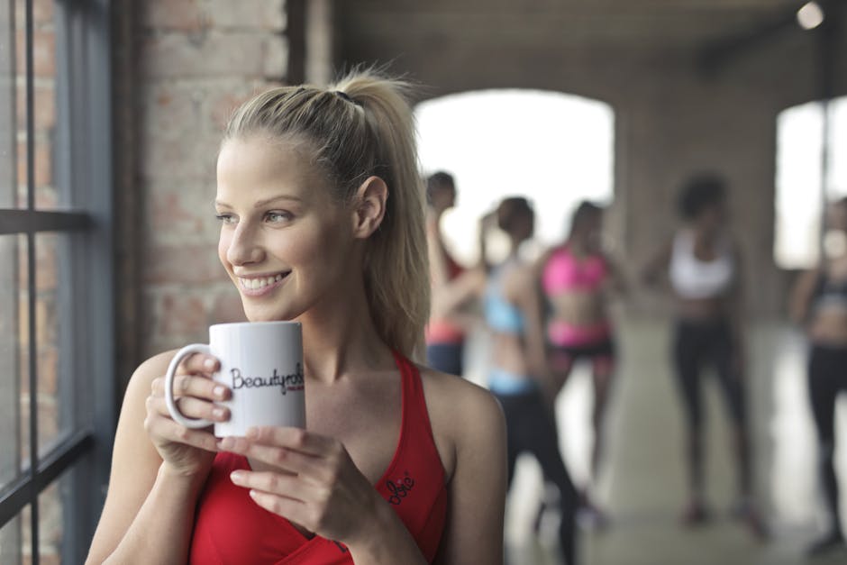 Kobieta w czerwonym topie trzyma biały kubek przy oknie w sali fitness.